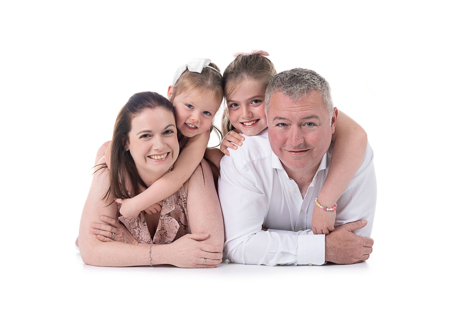A family lies together on the floor in white clothing against a white background for a portrait session.