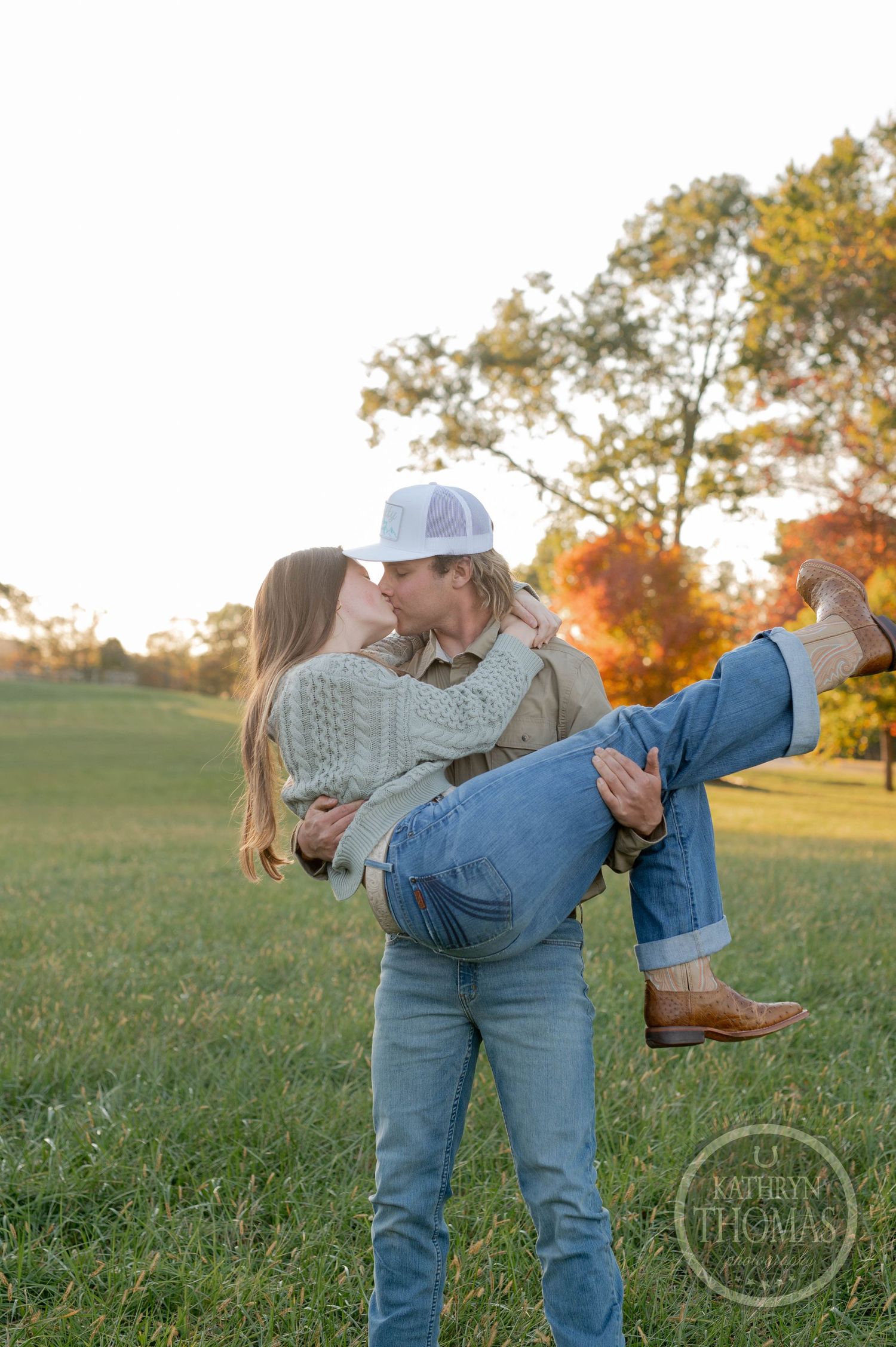 A playful moment captured as someone lifts their partner in front of autumn trees at sunset.