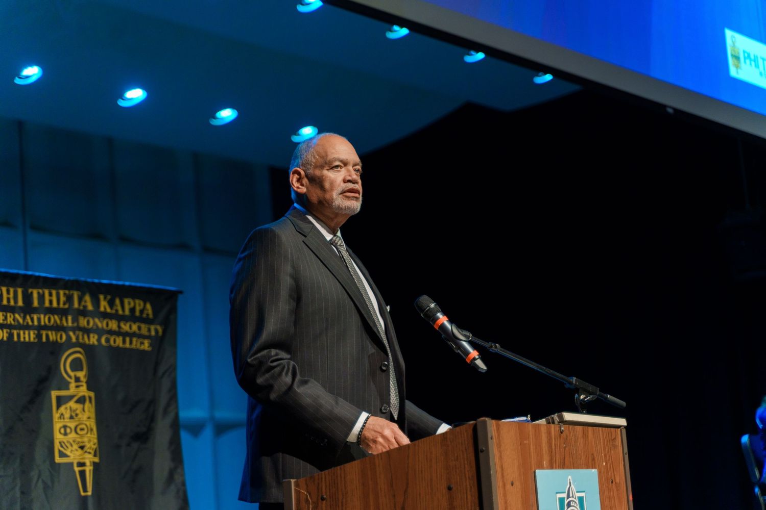 A speaker addresses an audience from behind a podium at a formal event with blue lighting.
