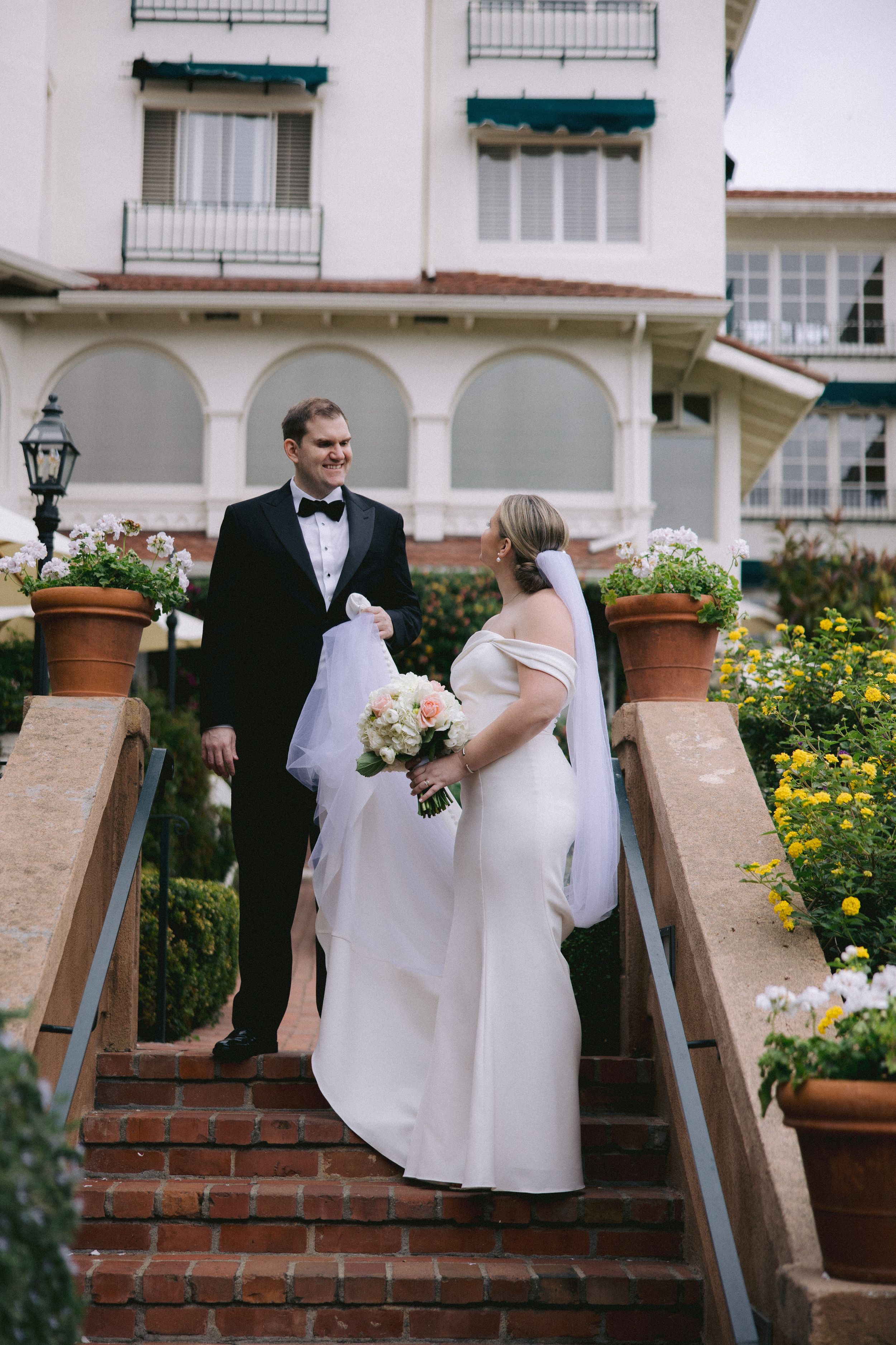 Wedding couple stands together on elegant brick steps surrounded by potted flowers and arched white architecture.