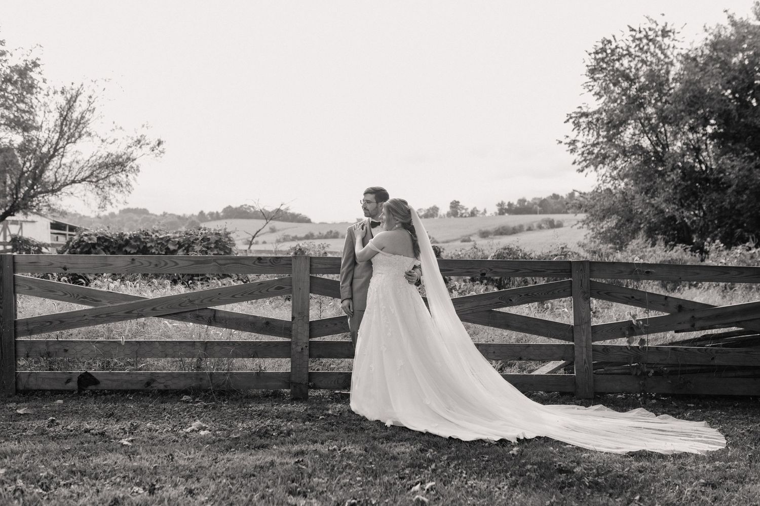A black and white wedding portrait captured against rustic wooden fences in a countryside setting.