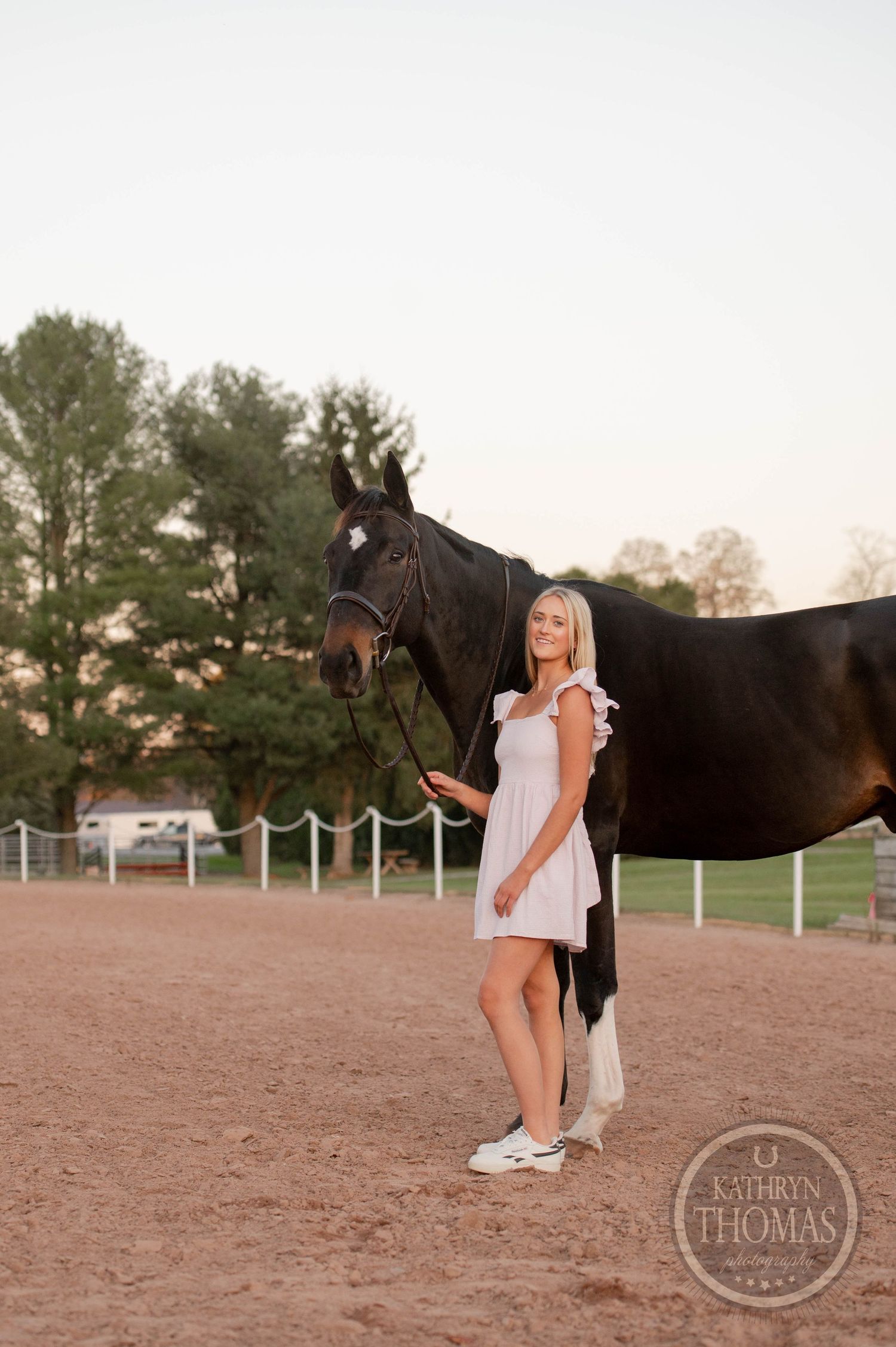 A person in a white dress stands beside a dark horse in a sandy arena.