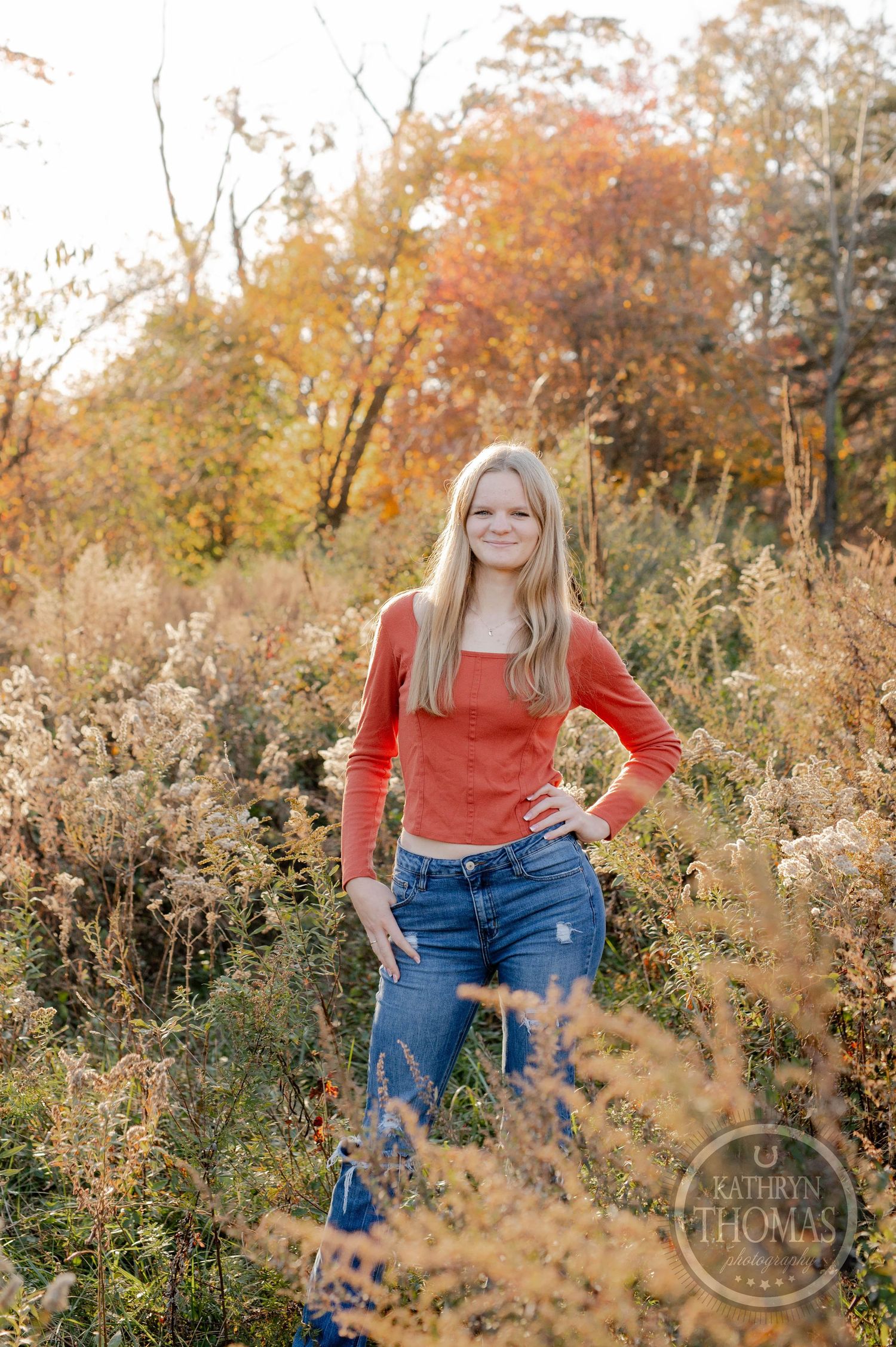 Standing portrait in rusty orange sweater and jeans among fall wildflowers at sunset.