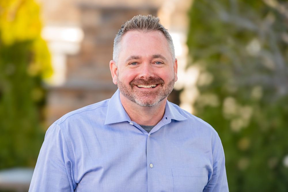 Professional branding headshot of person in blue button-down shirt against blurred outdoor background.