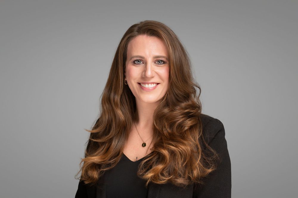 Professional headshot portrait of person with long brown wavy hair wearing black blazer against gray background.