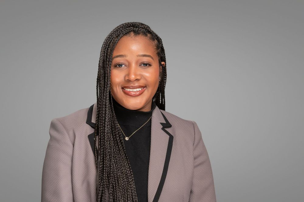 Professional headshot of a person with long braids wearing a mauve blazer and black turtleneck against a gray background.
