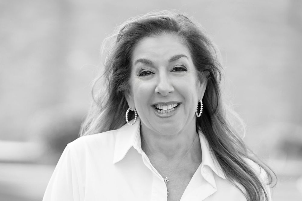 Black and white professional headshot portrait of a smiling woman with long hair wearing a white blouse and hoop earrings.