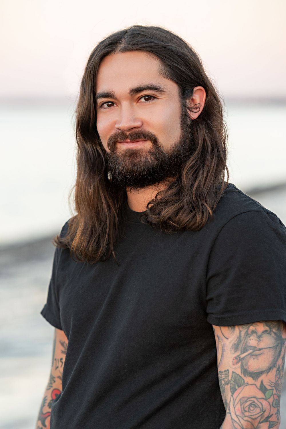 Person with long dark hair and full beard wearing a black t-shirt showing tattooed arms against a light waterfront background.