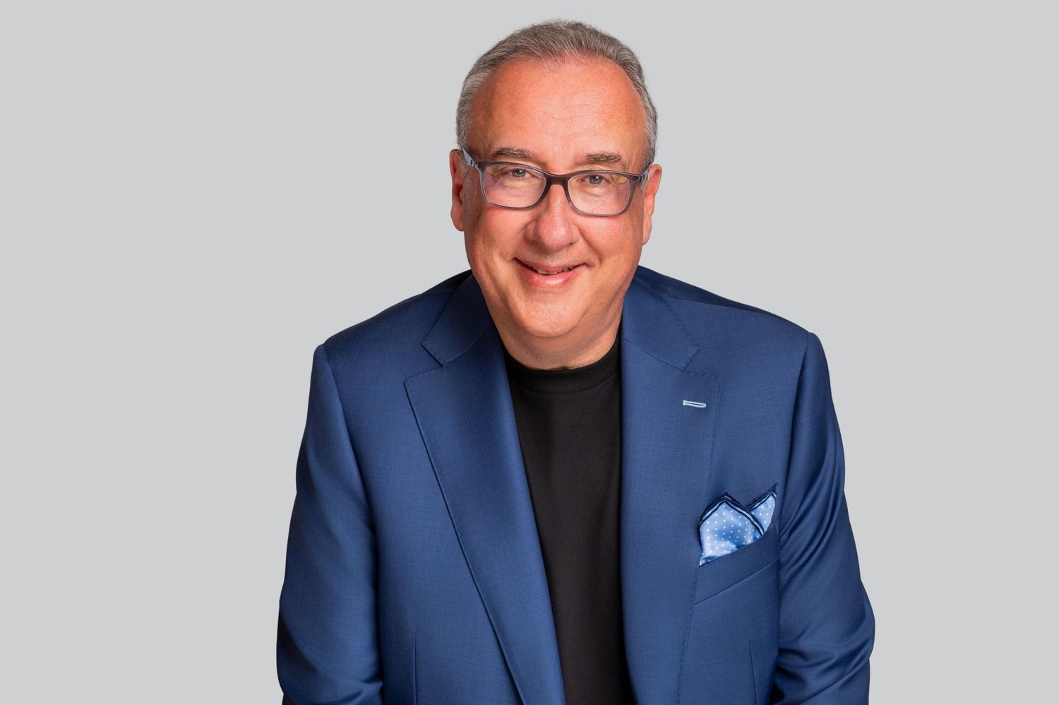 A man in a blue blazer and black shirt smiles warmly at the camera against a simple gray background.
