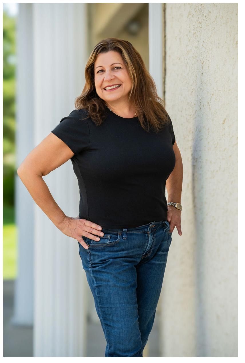 A smiling woman in a black t-shirt and jeans leans against a white column outdoors in a professional portrait photo.
