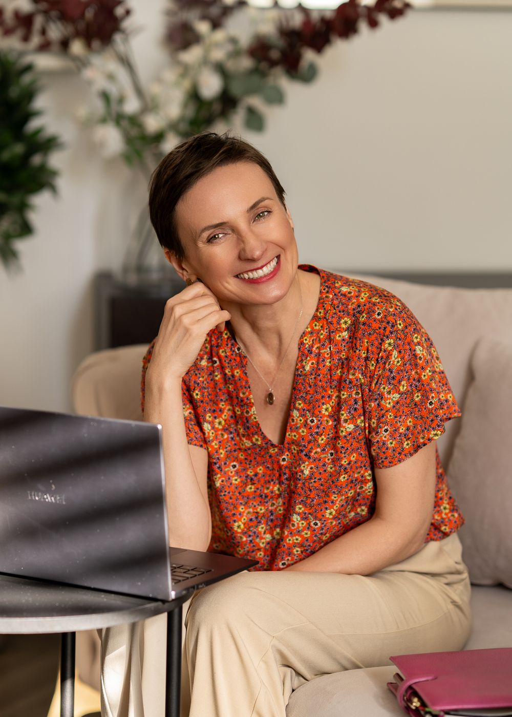 Cheerful portrait in floral top sitting at desk with laptop in home office setting.