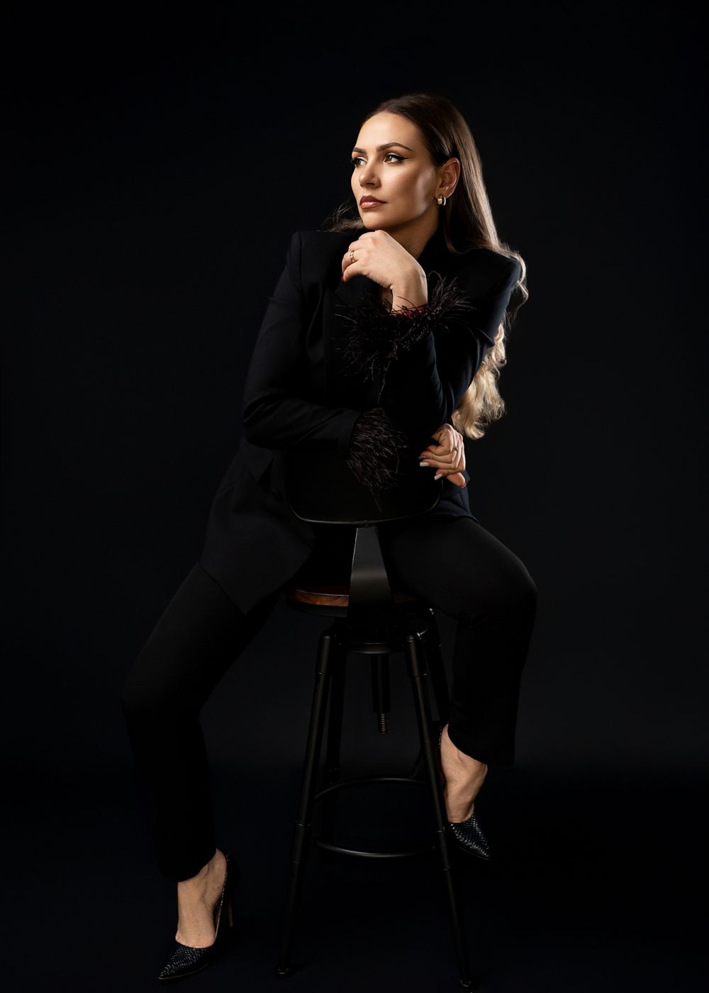 Fashion portrait of a woman in black outfit sitting on high stool against dark background.