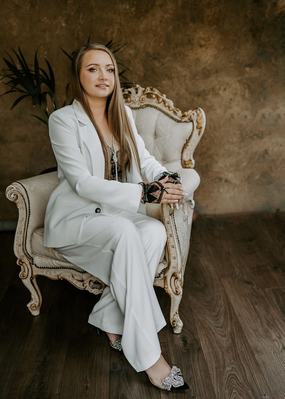 Professional wearing crisp white pantsuit poses elegantly in ornate vintage chair against textured brown wall.