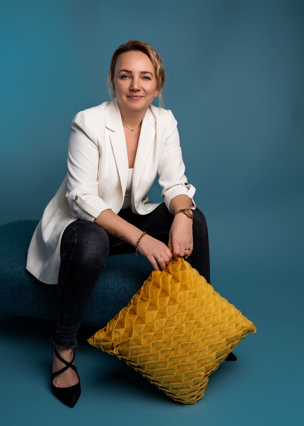 Professional woman in white blazer sits casually with yellow decorative pillow against blue backdrop.