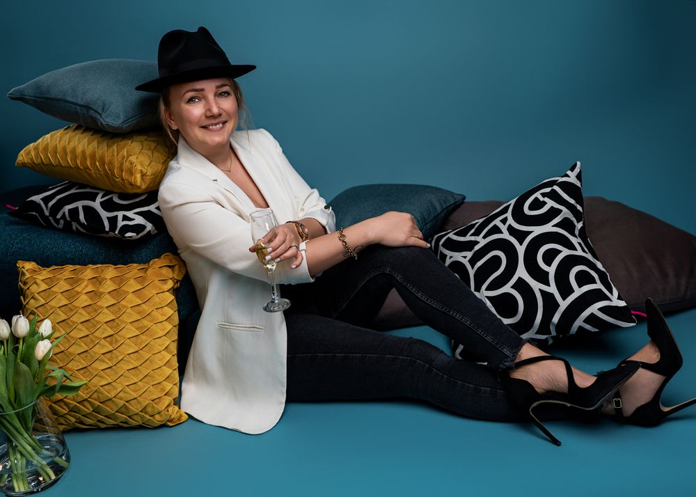 Stylish woman in white blazer and black hat reclines with yellow pillows on blue background.