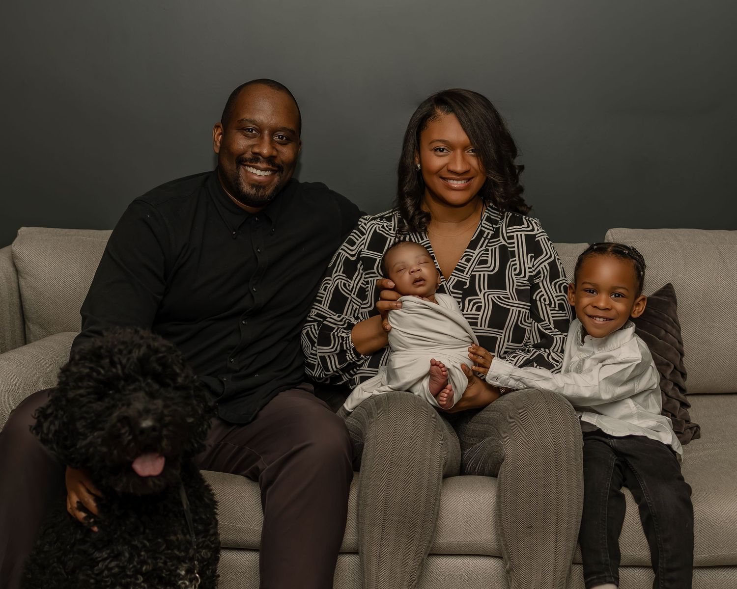 A family sitting together on a grey couch with their black dog, smiling warmly in a studio setting.