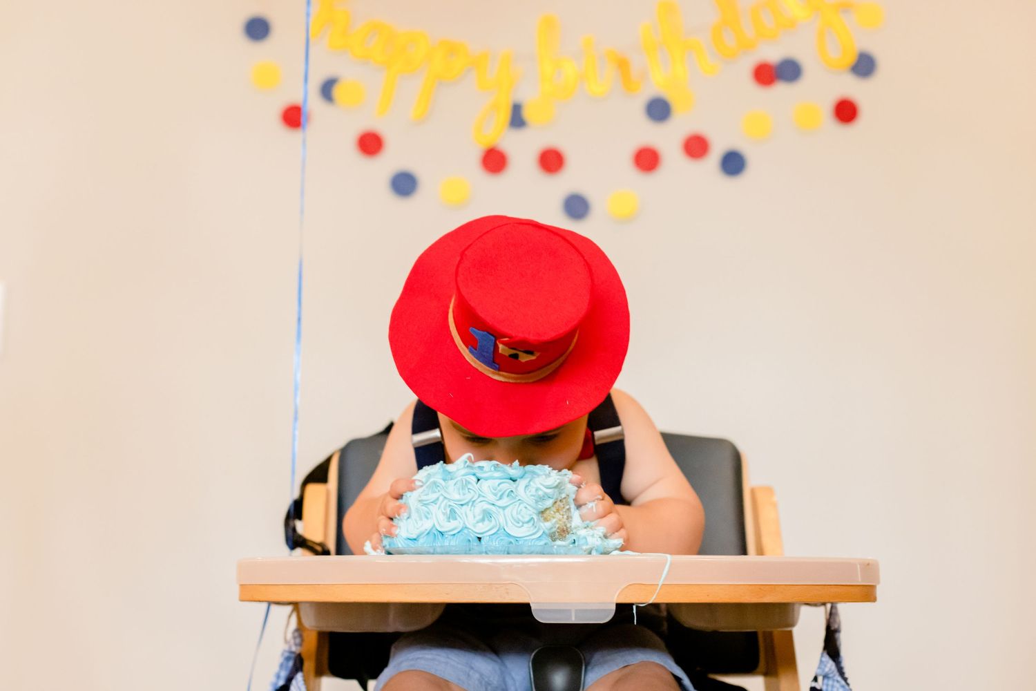 Baby wearing red hat sits in high chair under colorful birthday party decorations with polka dot banner.
