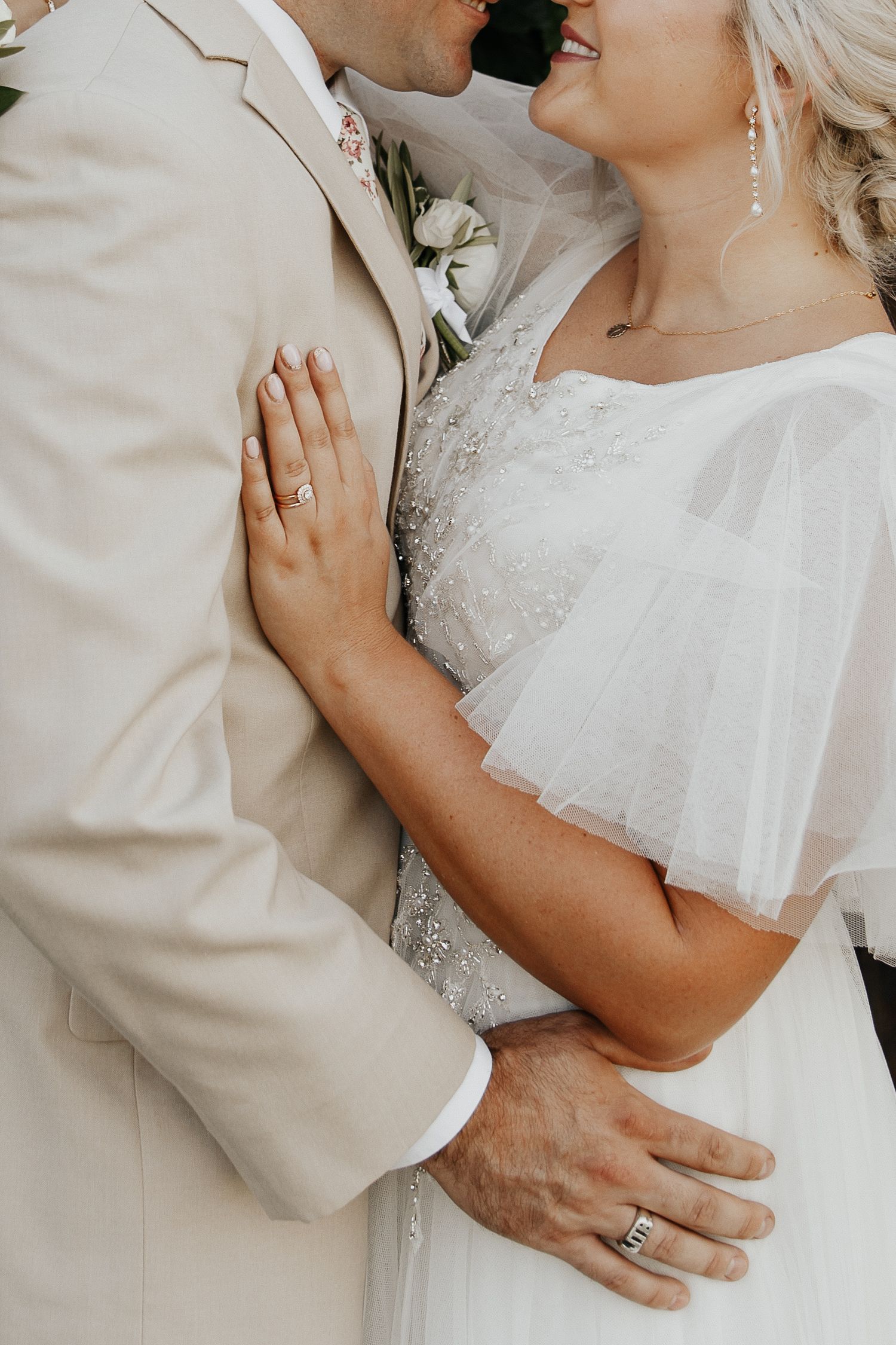 Close-up wedding photo of newlyweds embracing, with bride in white dress and groom in beige suit holding white flowers.