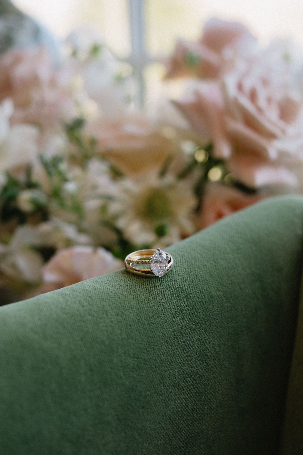 Diamond engagement ring placed on green velvet surface with soft pink flowers in background.