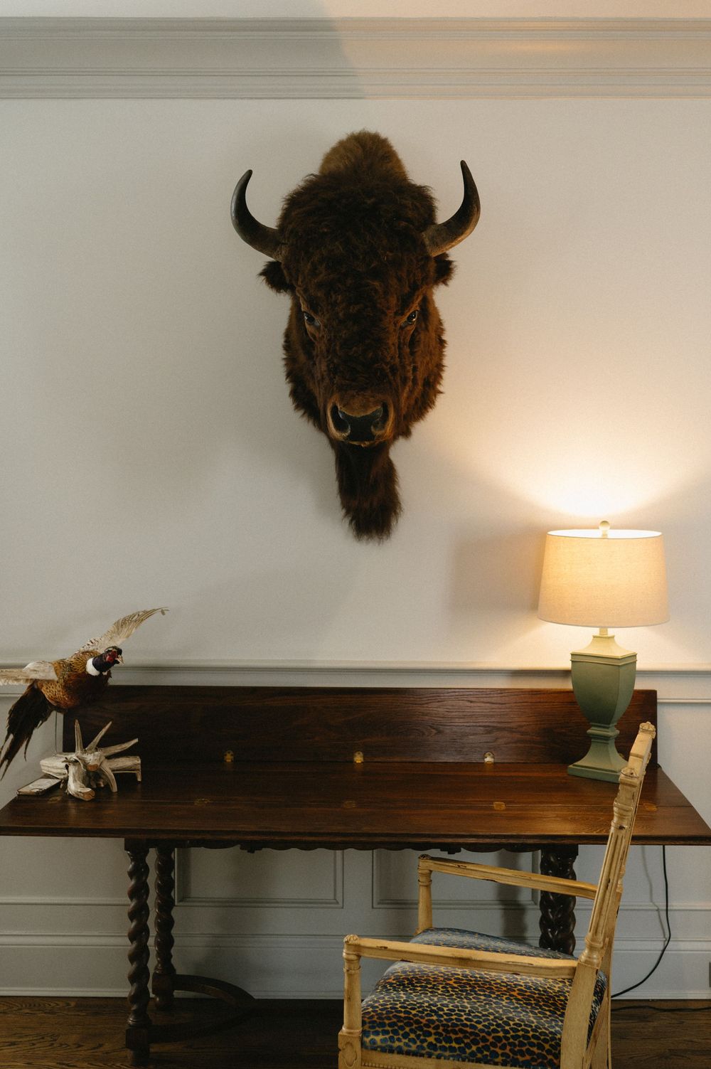Mounted buffalo head above a dark wooden console table with lamp and chair.