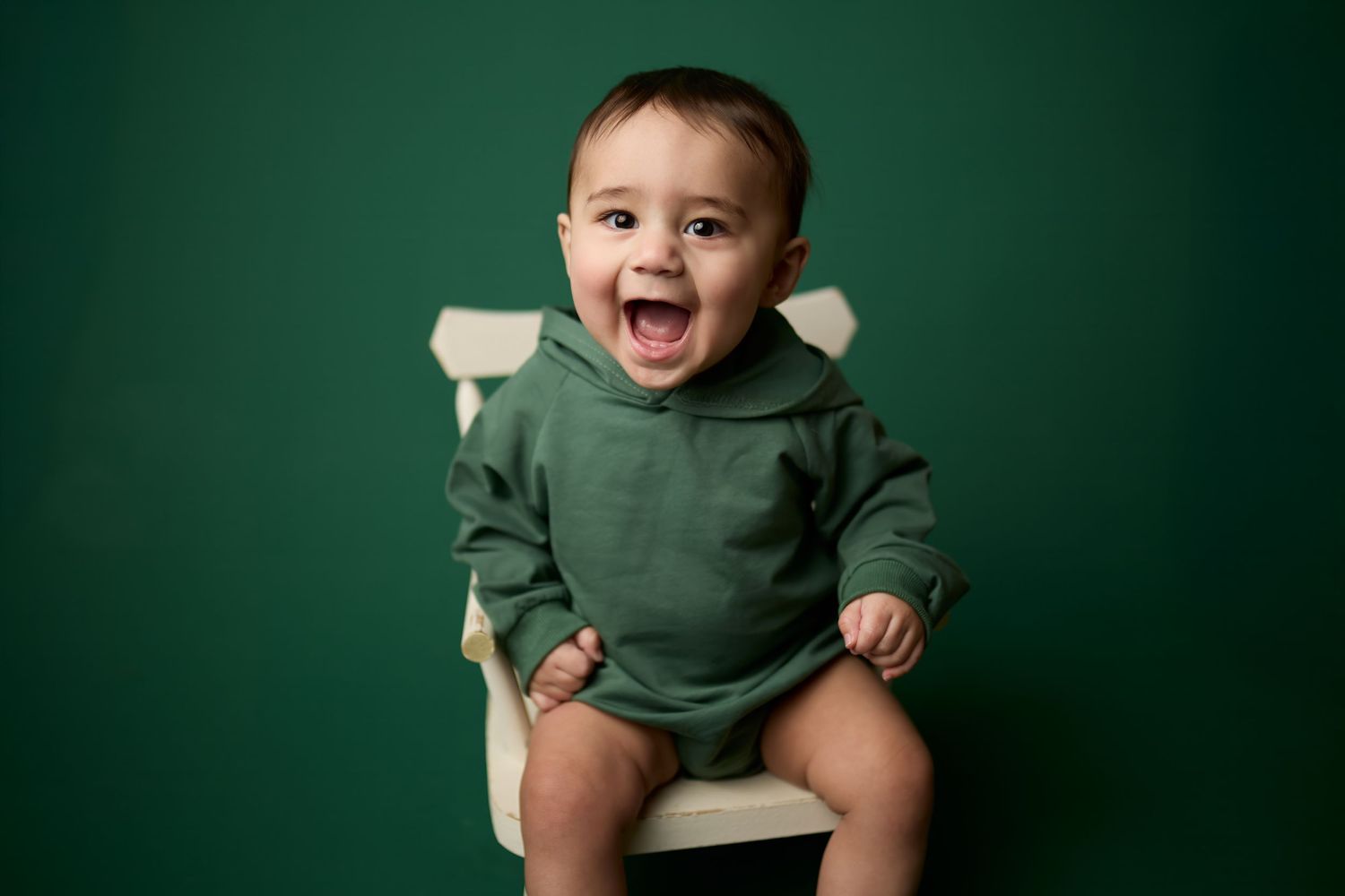 Baby wearing green hoodie laughs joyfully while sitting against emerald green backdrop during studio photo session.