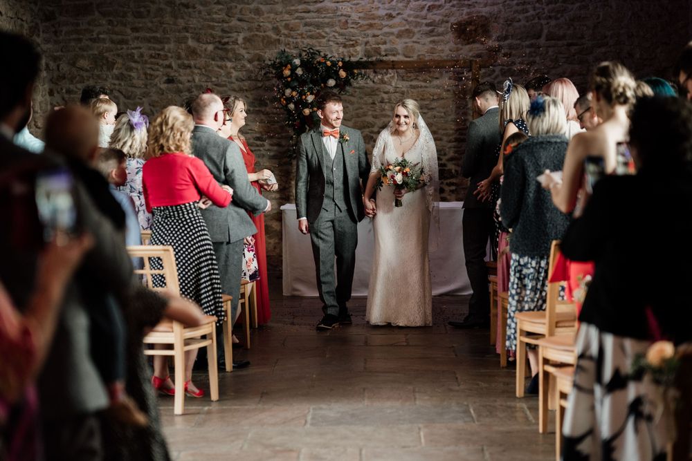 Wedding processional in stone-walled venue with guests seated on wooden chairs.