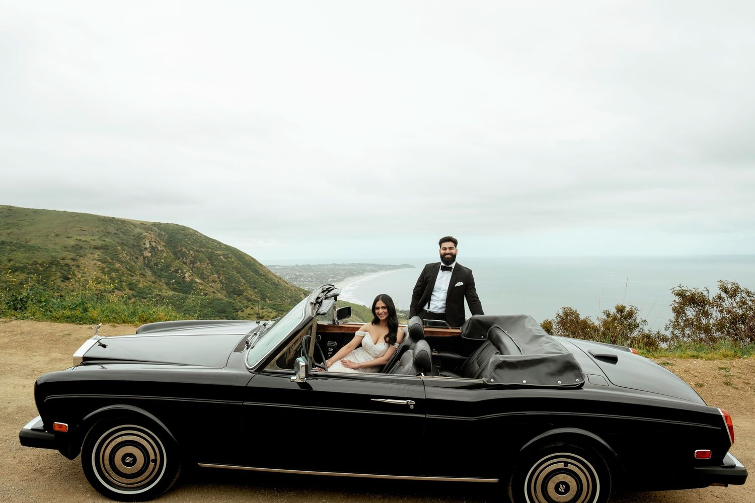 Classic black convertible car parked on a scenic mountain overlook along the coast on an overcast day.