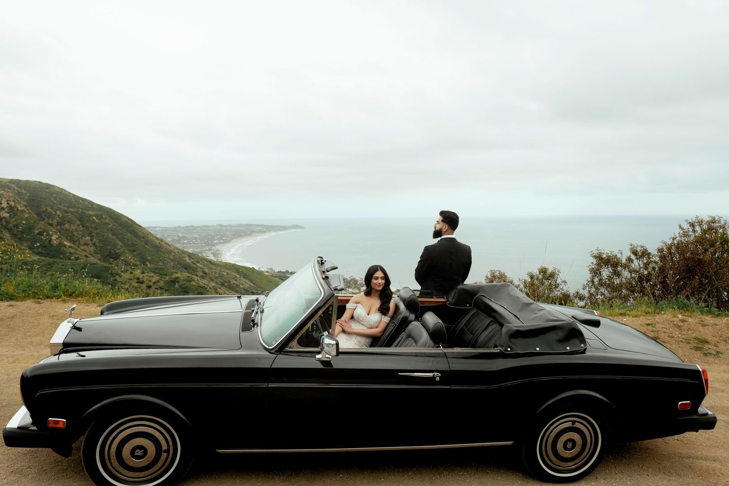 A vintage black convertible car parked along a scenic coastal overlook with rolling hills and ocean views.