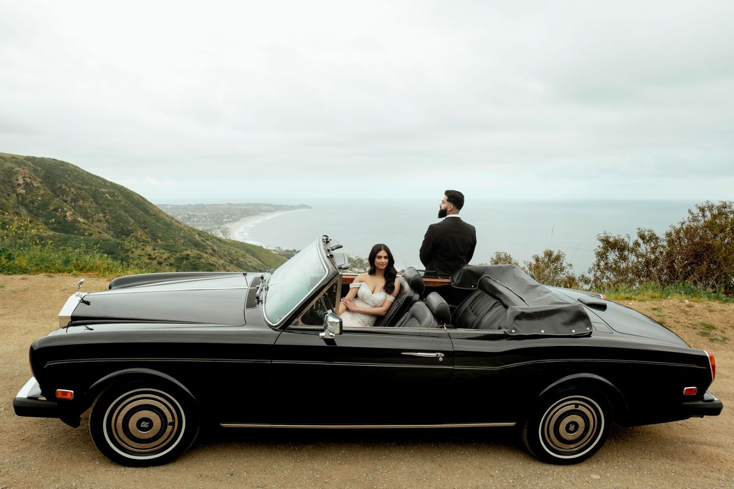 Classic black convertible car driving along a scenic coastal mountain road on an overcast day.