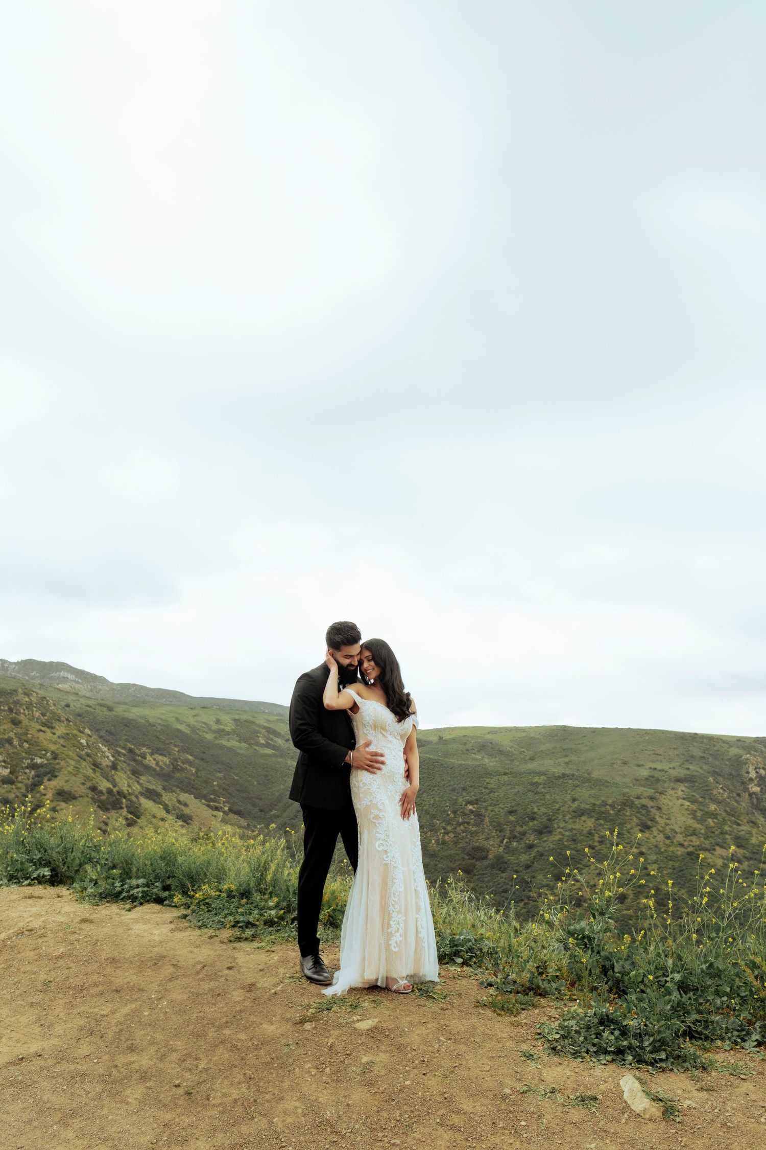A couple shares a romantic moment on a hilltop trail overlooking green coastal hills under an overcast sky.