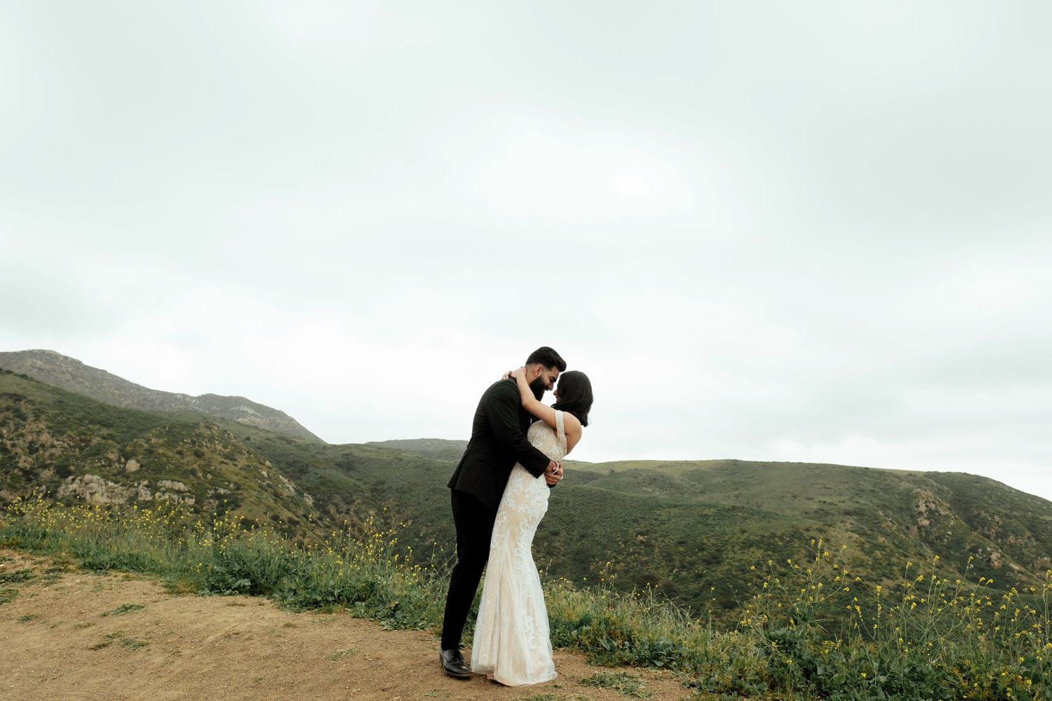 A romantic couple in wedding attire embraces atop a scenic hillside overlooking green rolling mountains on an overcast day.