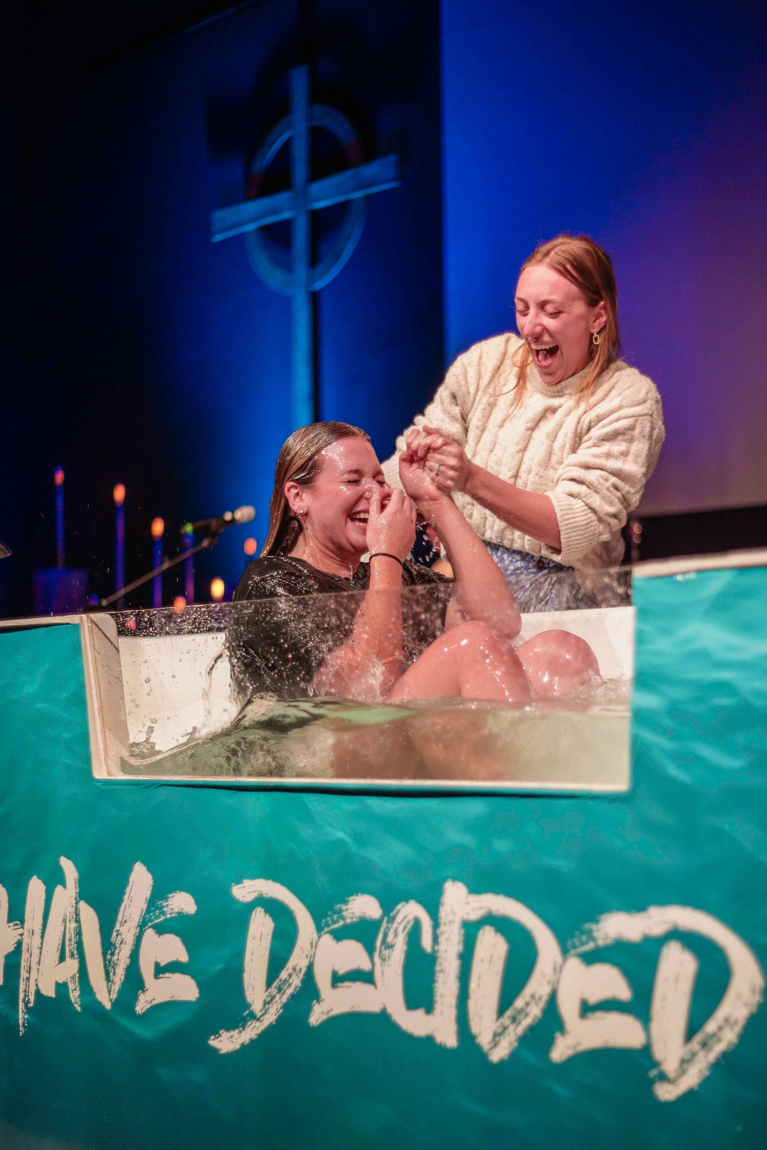 A person being baptized in a clear tank during a church ceremony with a cross symbol illuminated in the background.