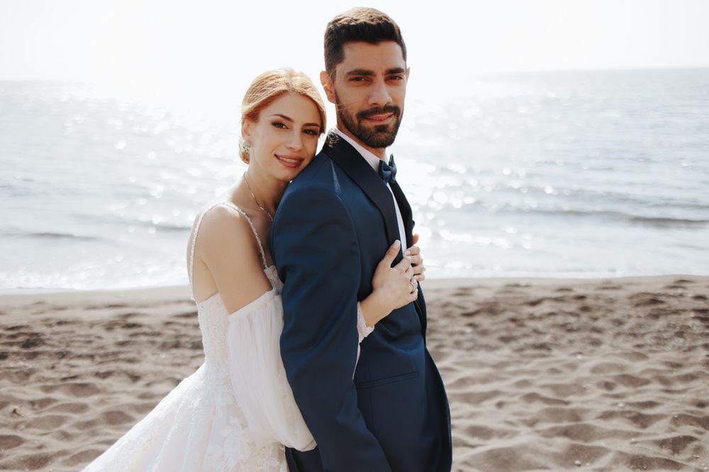 A couple in wedding attire poses together on a sunny beach with ocean waves in the background.