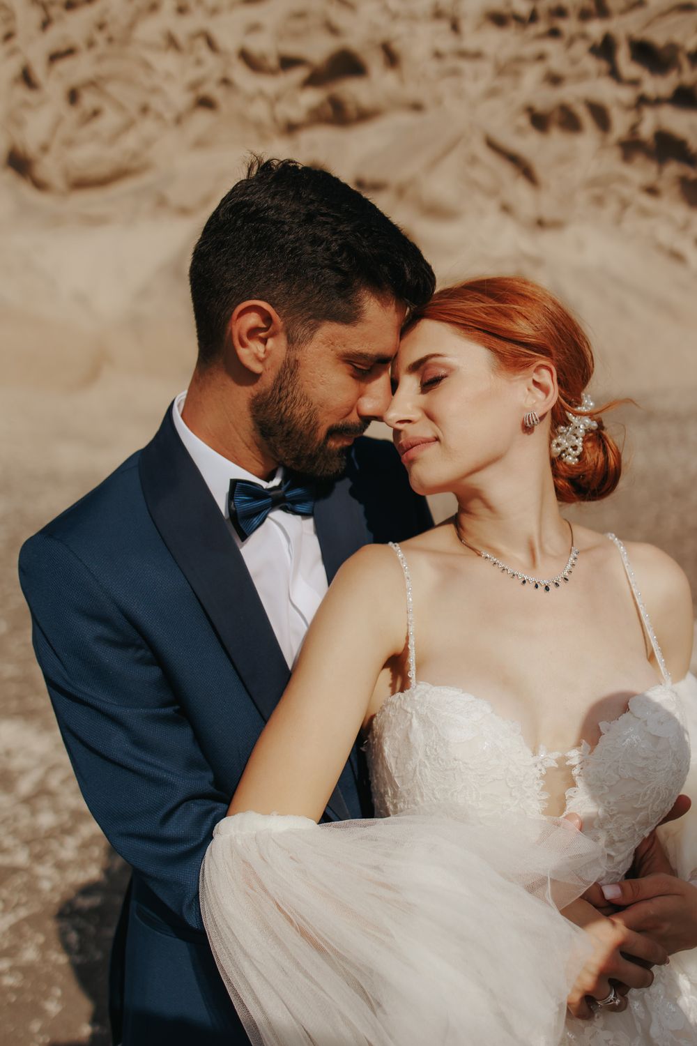 A romantic wedding portrait with a groom in navy suit and bride in white dress embracing against a sepia-toned background.