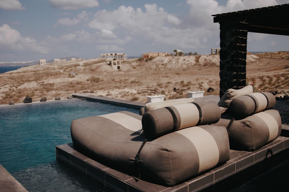 Beige cushioned lounge seating overlooks a stunning coastal view with blue water and rocky terrain.