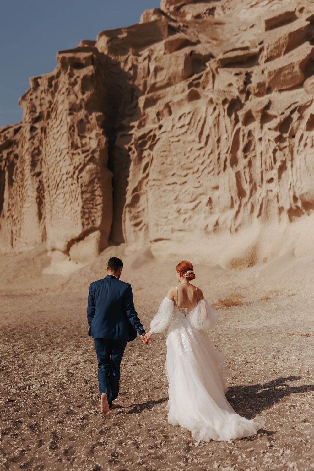 A couple in wedding attire walks hand in hand along a desert path with towering red rock cliffs in the background.
