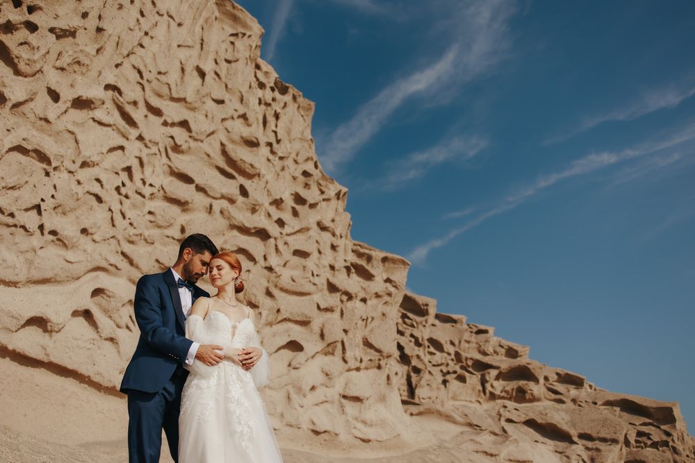 Two people in wedding attire embrace near a weathered desert cliff formation under a bright blue sky.