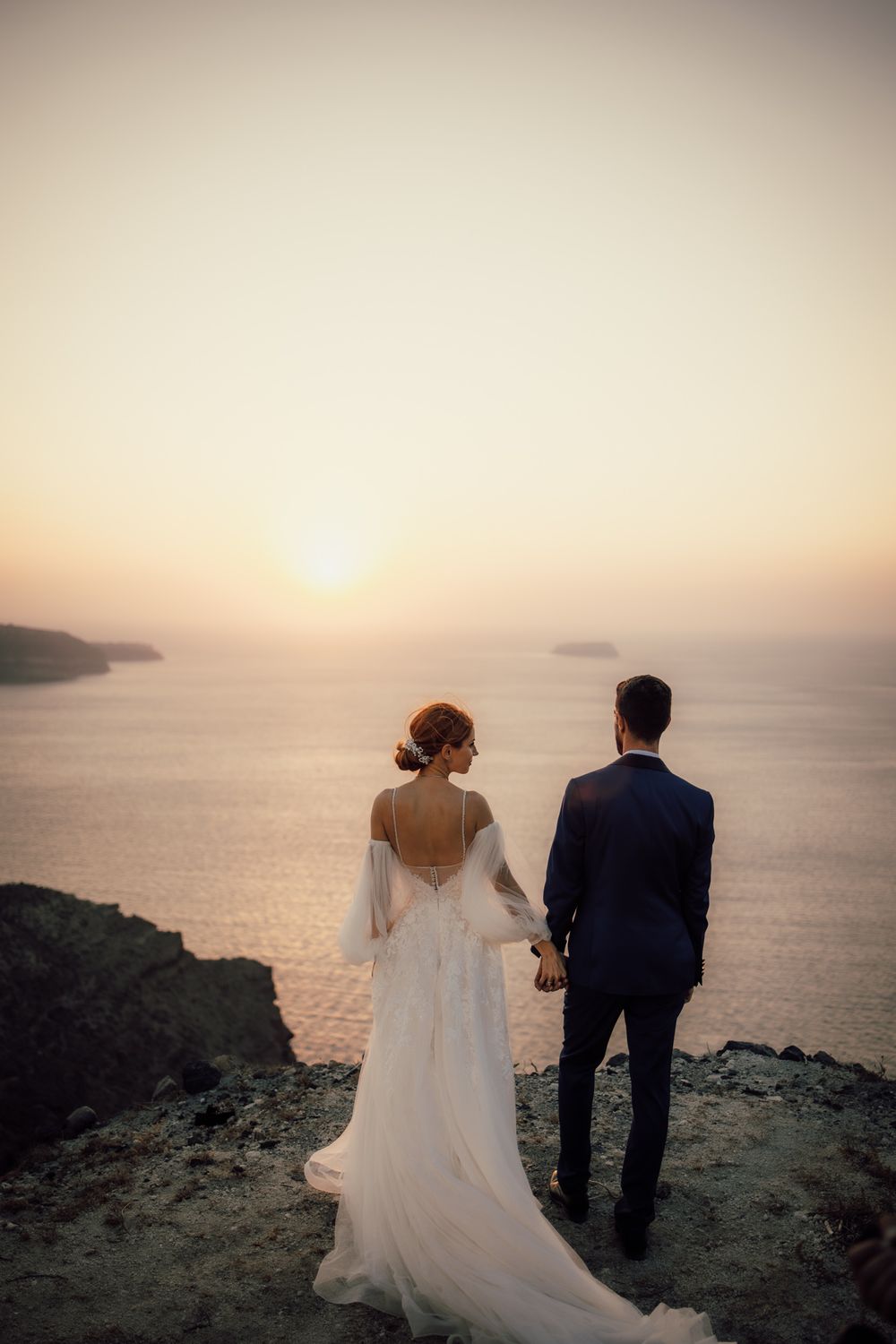 Newlywed couple holding hands while watching sunset over ocean from dramatic cliff edge in wedding attire.
