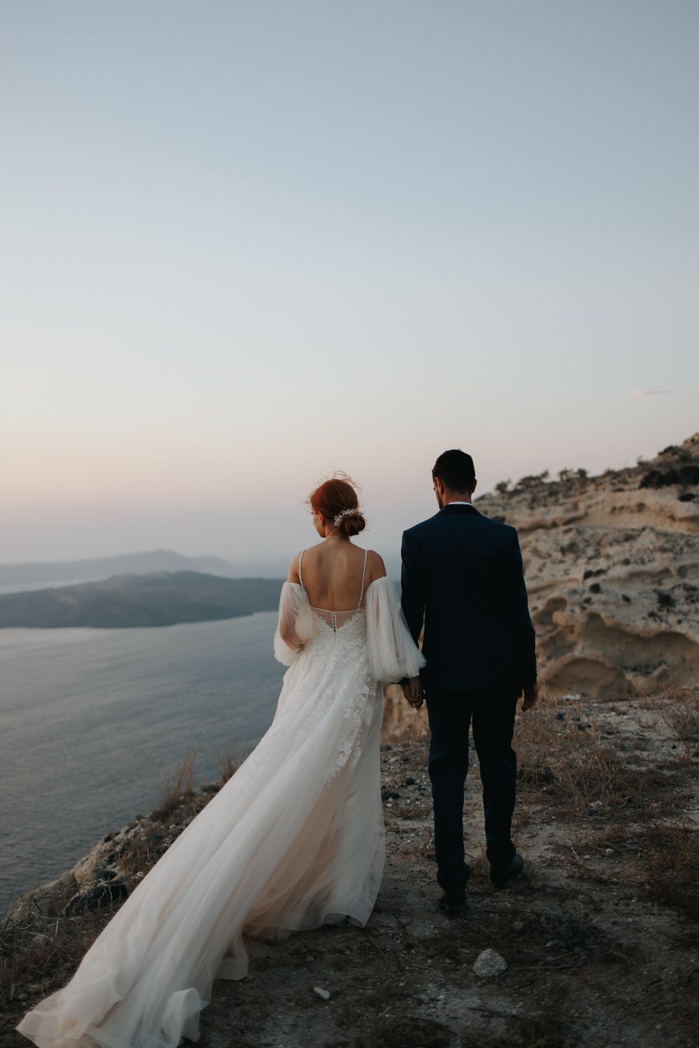 Couple in wedding attire looking at scenic mountain view over water during sunset in Santorini.