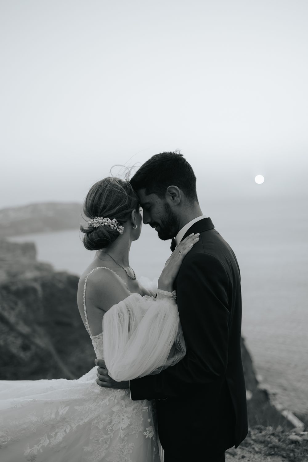 A romantic black and white wedding photo of a couple embracing on a misty coastal cliff with a full moon in the background.