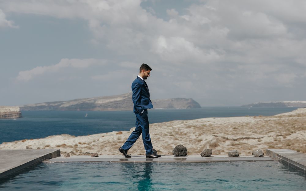 Person in blue suit walks along edge of infinity pool with dramatic coastal landscape in background.