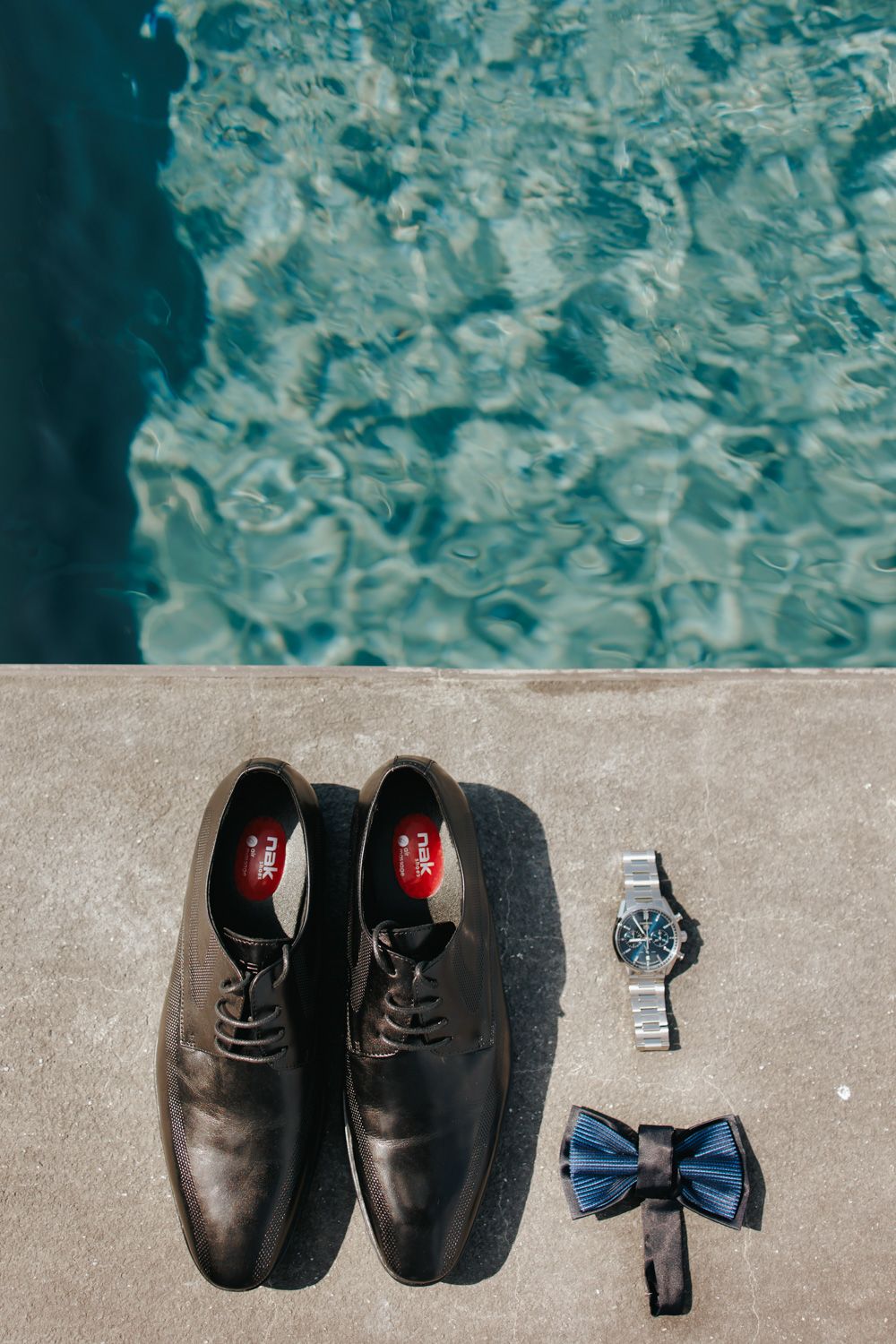 Black leather dress shoes arranged with a blue bow tie and silver watch on a concrete surface beside turquoise water.