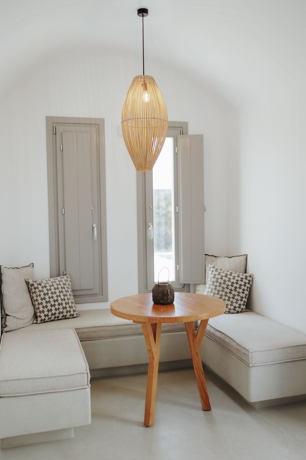 Modern minimalist seating area with woven pendant light, round wooden table, and built-in cream benches with patterned pillows.