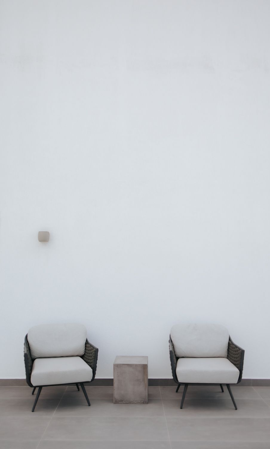 Two minimalist white armchairs with dark frames flank a concrete side table against a light gray wall.