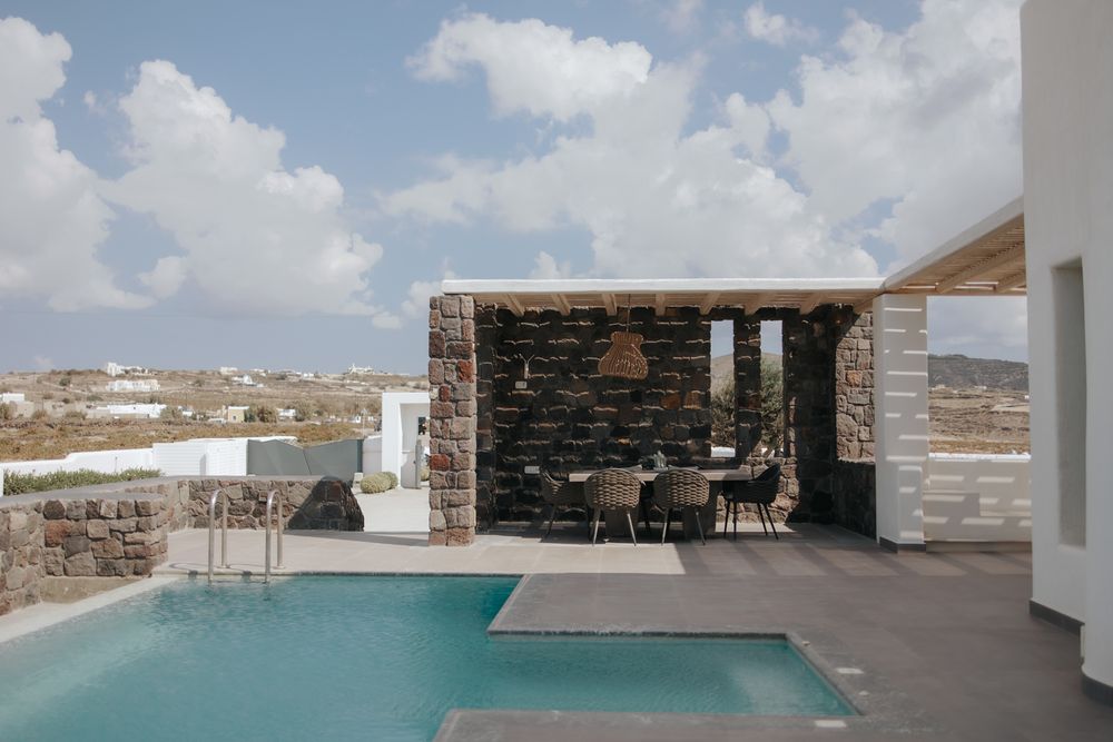 Luxurious villa patio with infinity pool and stone-clad pergola overlooking Mediterranean landscape under blue sky.