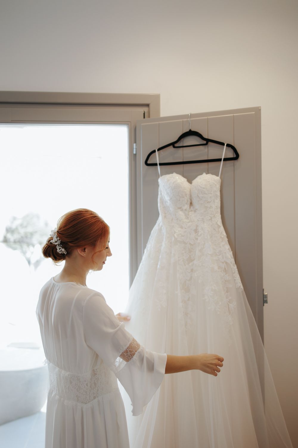 A white bridal gown with lace details hangs on display while someone in a white robe examines the dress near a window.