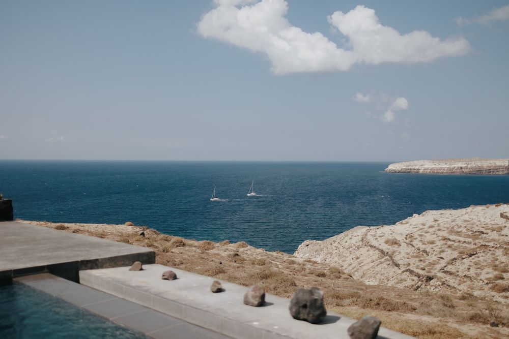 Modern terrace overlooking a dramatic Mediterranean coastline with rocky cliffs and azure blue water under scattered clouds.
