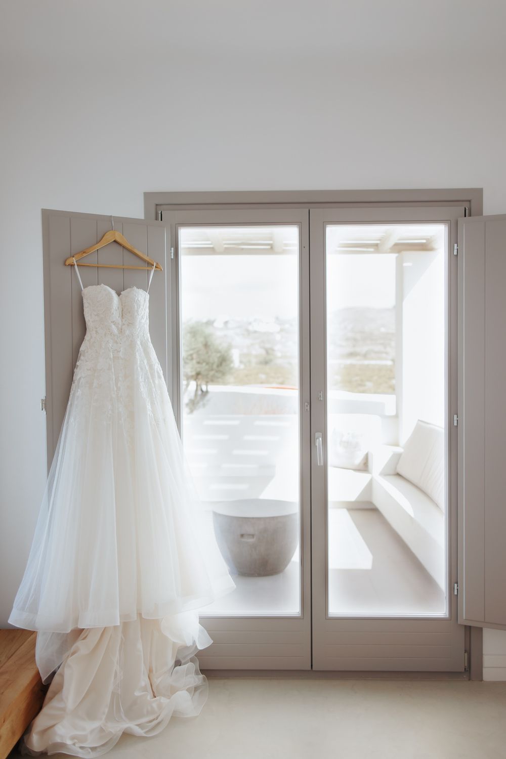 A white wedding dress with lace details and tulle skirt hangs elegantly by a sunlit French door window.
