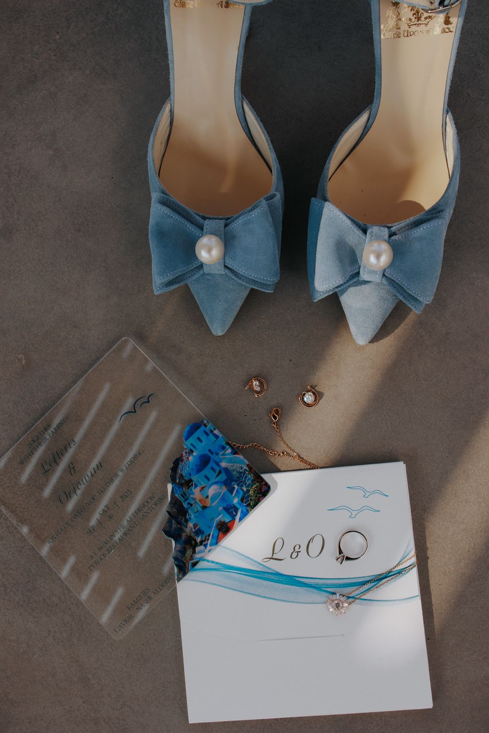 Elegant light blue suede bridal shoes with pearl bows displayed alongside wedding accessories and invitation on gray surface.