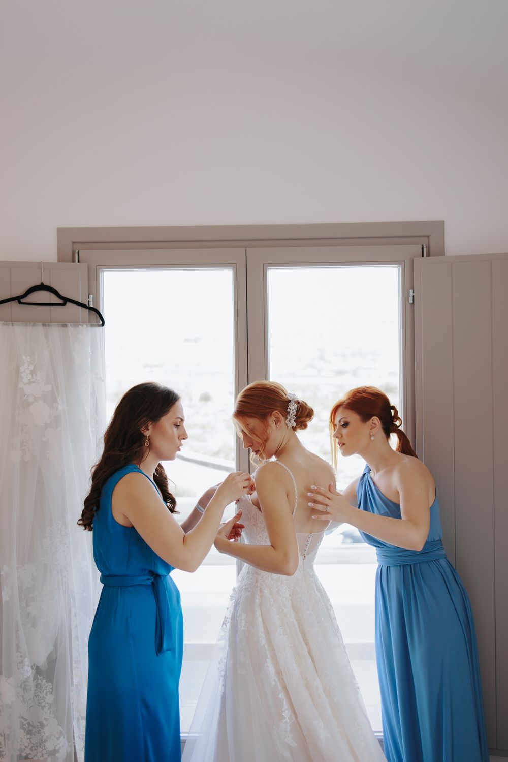 Three women in blue and white dresses help with wedding preparations by a bright window.